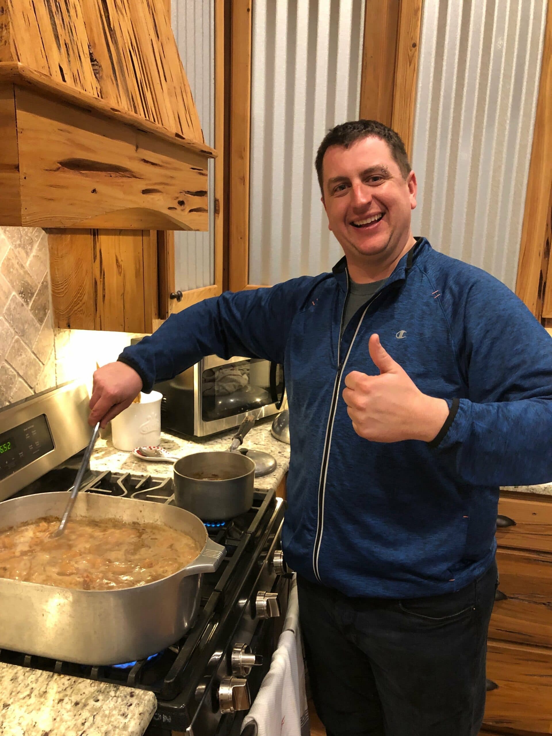 A smiling man stirs a pot of food on the stove while giving a thumbs up.