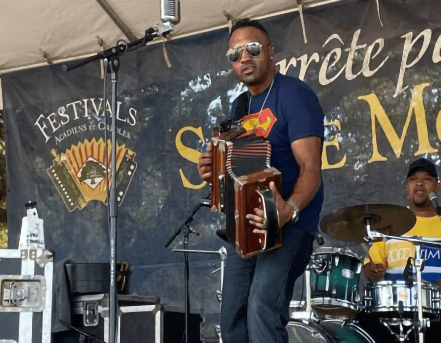 A musician plays the accordion on stage at a festival, with a drummer visible in the background.