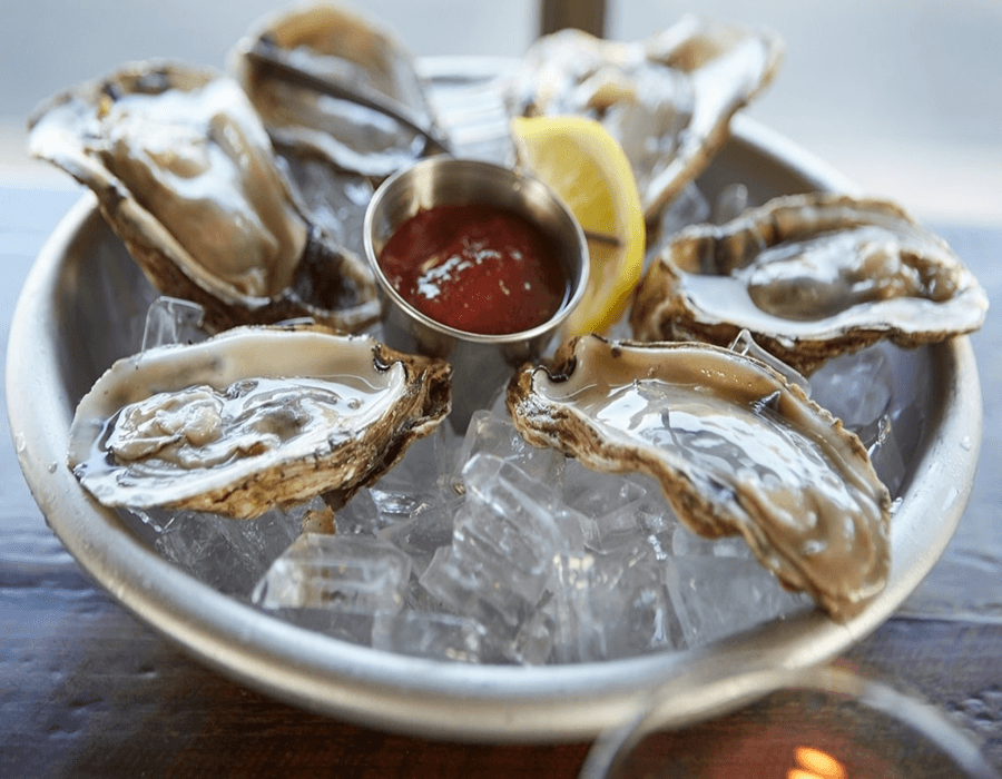 A silver platter holds several fresh oysters on ice, accompanied by a lemon wedge and cocktail sauce.