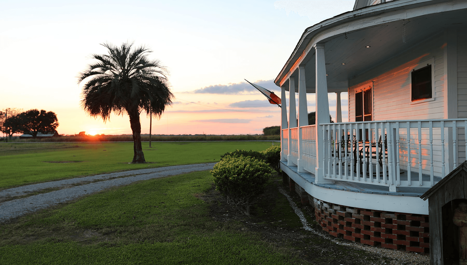 A white porch with rocking chairs overlooks a sunset-lit landscape featuring palm trees and grassy fields.