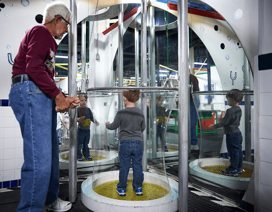 An elderly man operates a science exhibit while a young boy stands inside a transparent chamber.