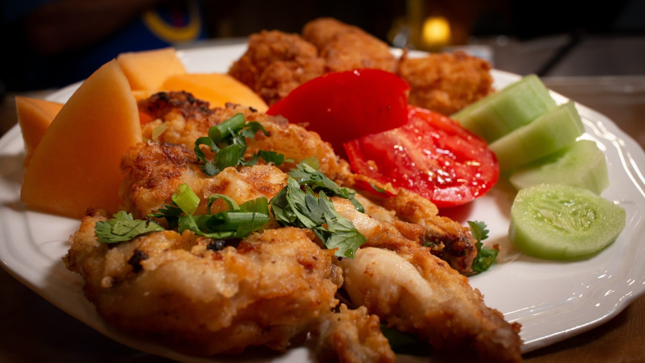 A plate of fried fish and chicken accompanied by slices of tomato, cucumber, and cantaloupe.