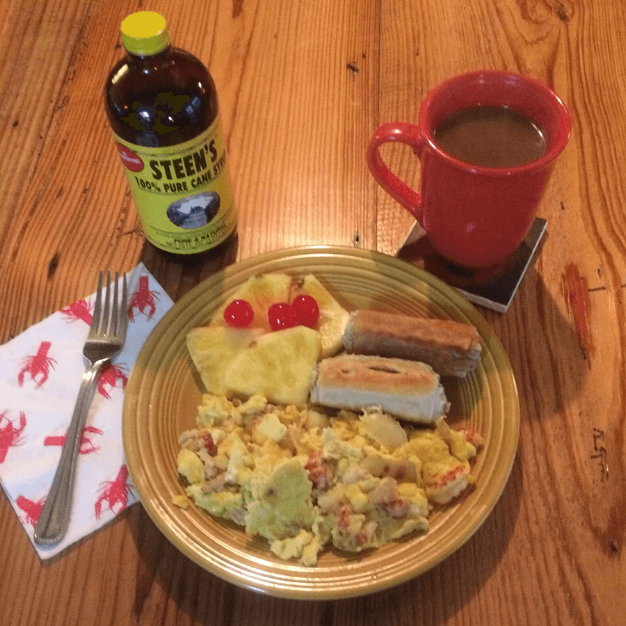 A hearty breakfast plate featuring scrambled eggs with crawfish, sausage rolls, pineapple, maraschino cherries, a bottle of cane syrup, and a cup of coffee.