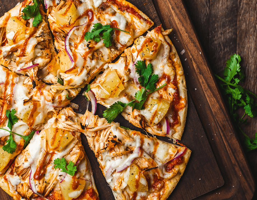 A delicious pizza topped with chicken, pineapple, red onions, and cilantro drizzled with barbecue sauce, served on a wooden board.