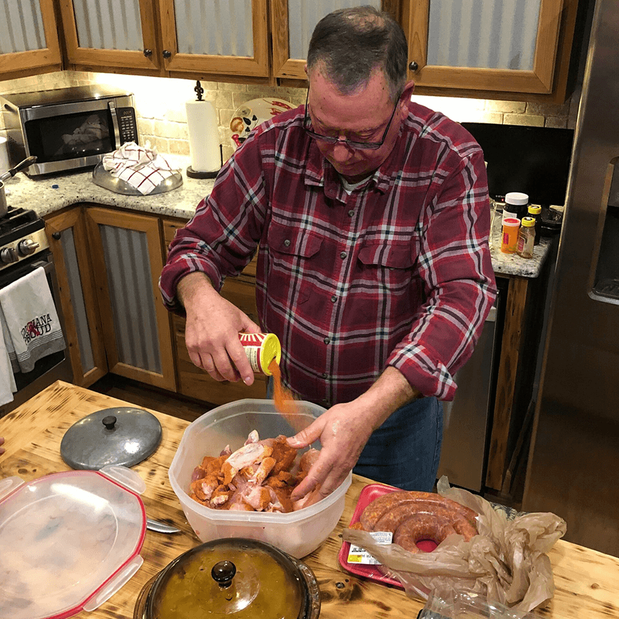 A man in a plaid shirt pours spices over raw chicken parts in a kitchen.