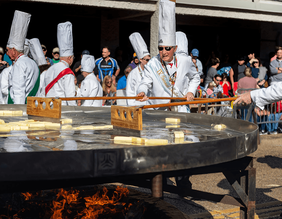 Chefs in white uniforms cook large blocks of food in a giant pan over an open flame at a festival.