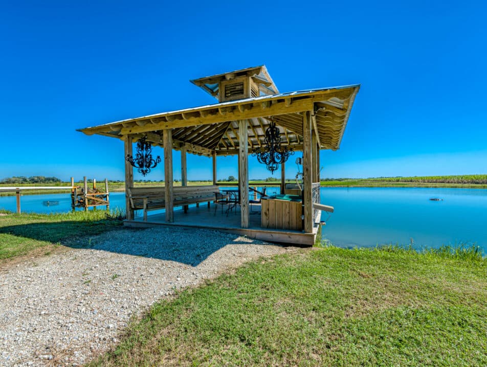 A wooden gazebo with a thatched roof overlooking a calm, blue pond under a clear sky.